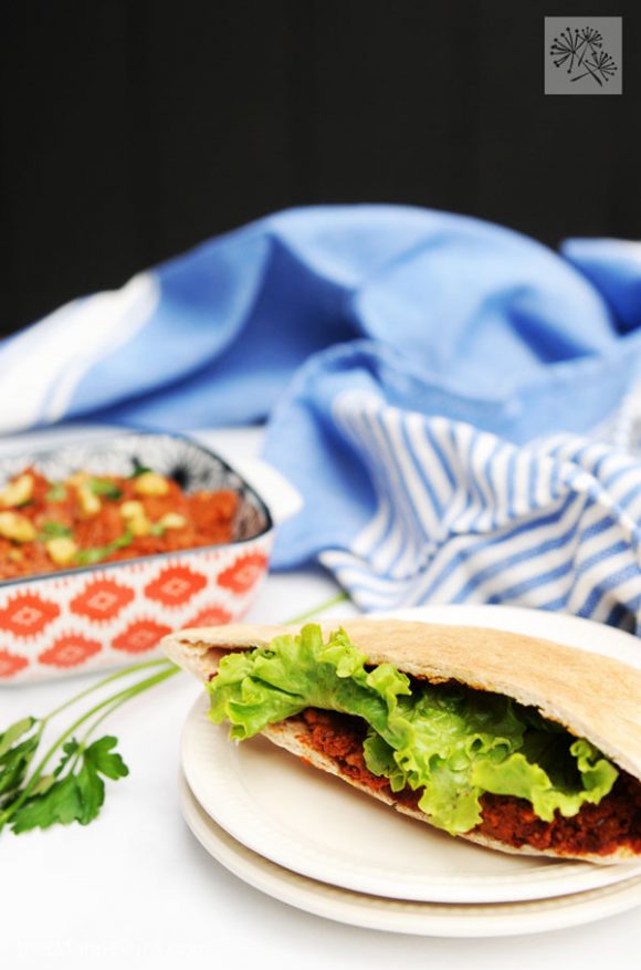 close up of a pita sandwich with a dish of muhammara in the background, along with a blue napkin and a black wall