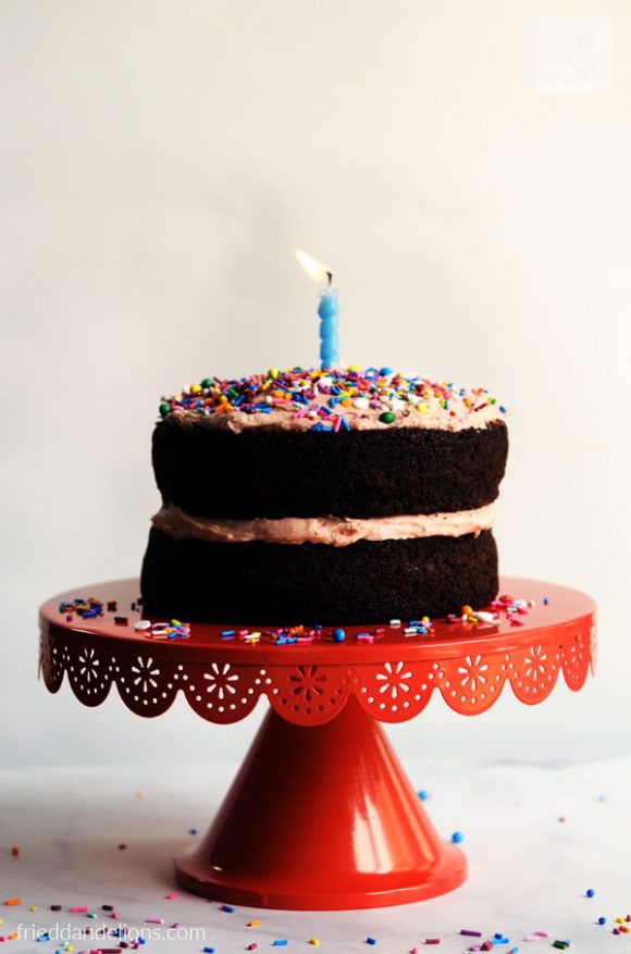 a vegan chocolate birthday cake on a red cake stand with a blue birthday candle burning