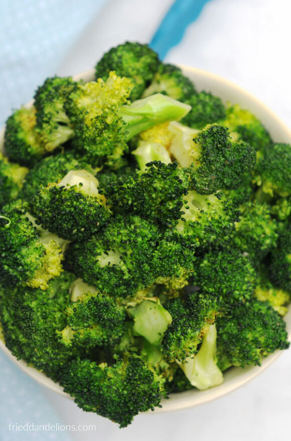 overhead view of bowl of air fried broccoli with blue tongs in background