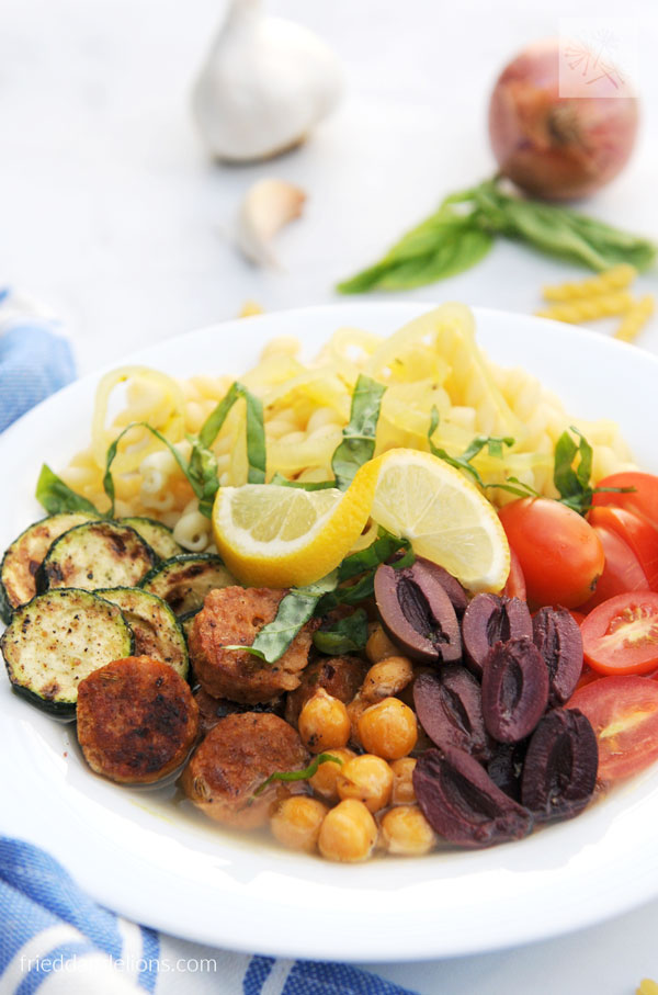 pasta power bowl with pasta, tomatoes, olives, chickpeas, sausage, zucchini, and lemon with blue napkin