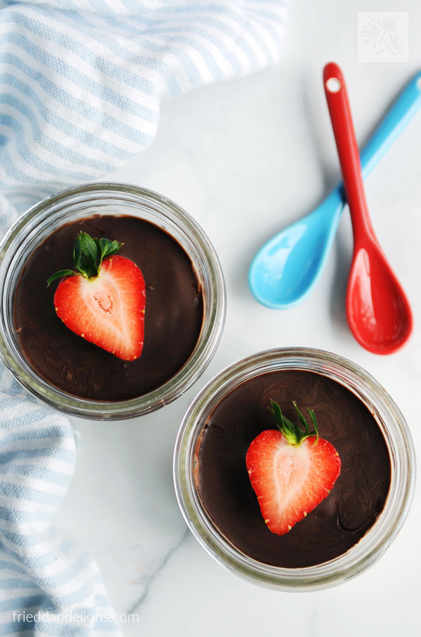 two vegan vanilla pudding cups with strawberry garnish and red and blue spoons crossed in the background