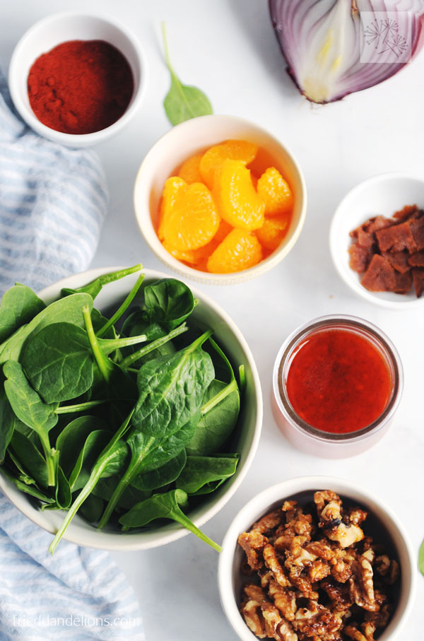 overhead shot of ingredients used to make spinach salad with bacon: oranges, paprika, onion, spinach, vegan bacon, candied walnuts