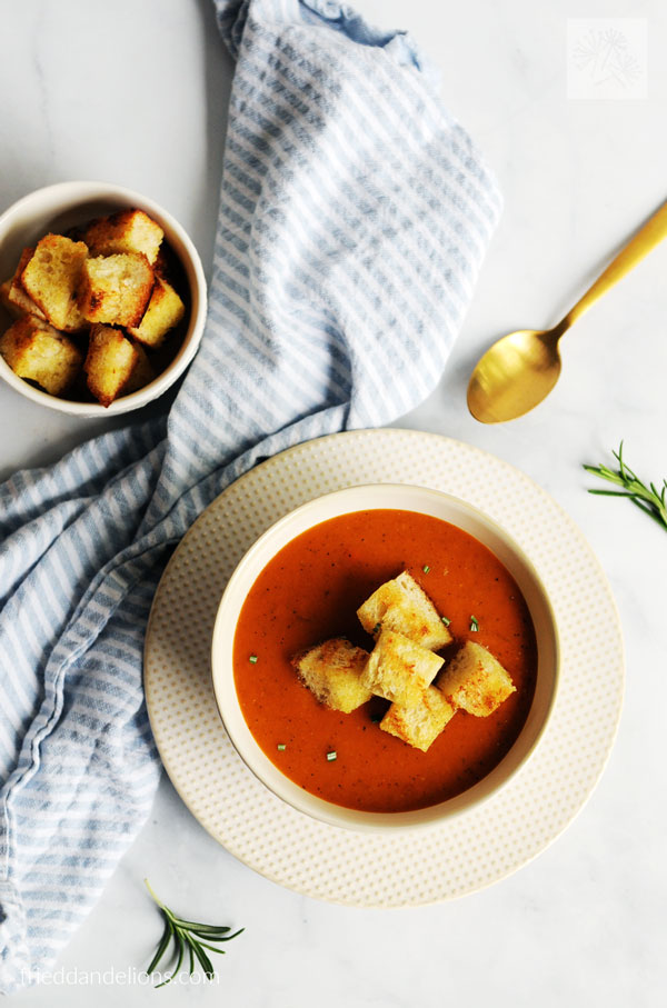 overhead view of bowl of vegan tomato soup with blue striped napkin, gold spoon, croutons in background