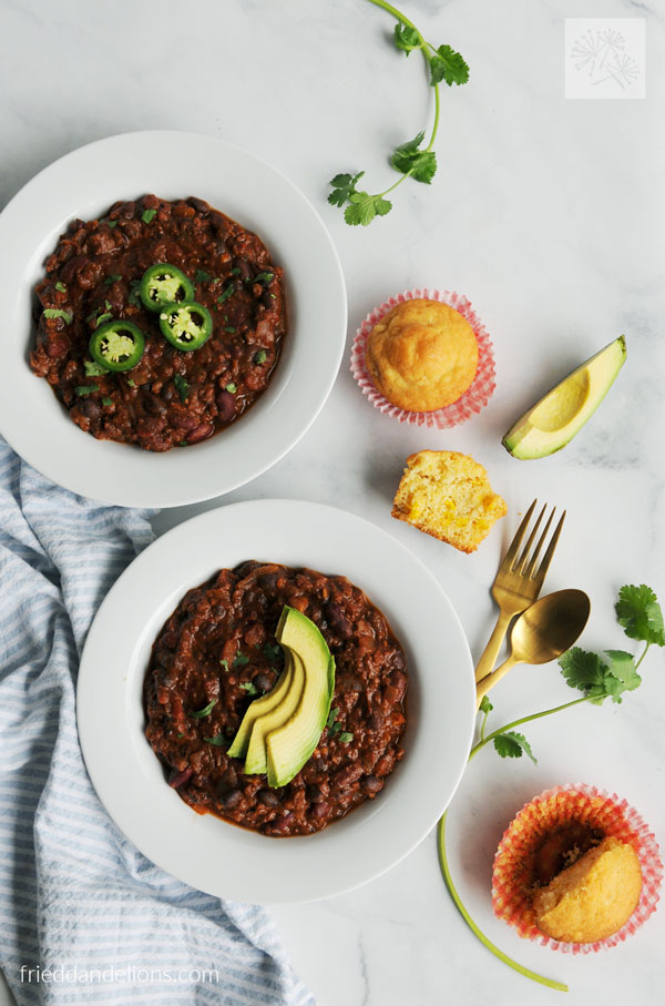 overhead view of two bowls of easy vegan chili with cornbread, cilantro, and avocado in background