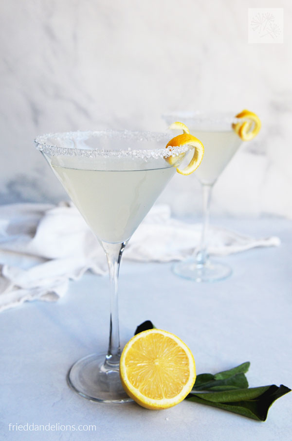 front view of two lemon drop cocktails with lemon in front and white dish towel in background