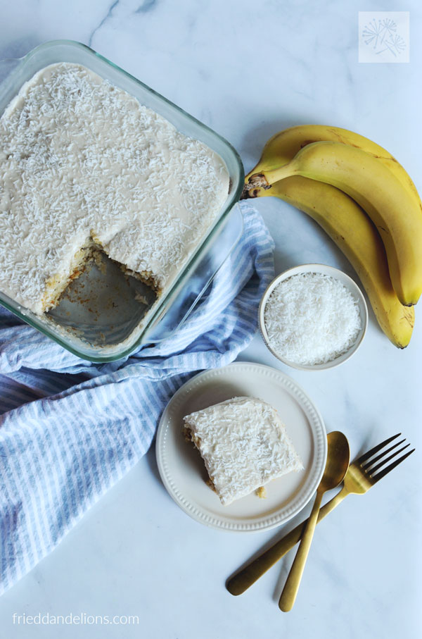 overhead view of banana coconut snack cake with one piece on plate, bananas, coconut, silverware in background